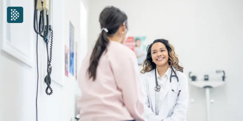 Médica em traje profissional demonstrando excelência organizacional durante consulta, conversando com paciente em ambiente de consultório médico moderno e bem equipado.