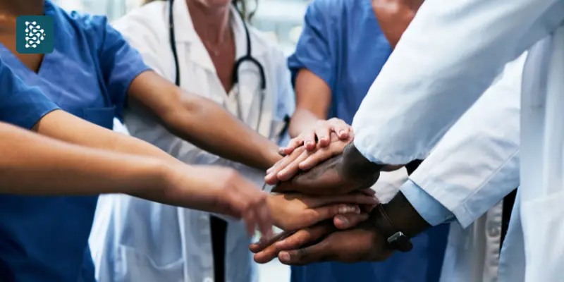 Médicos de jaleco branco unindo as mãos em um círculo, simbolizando trabalho em equipe e Excelência Organizacional na área da saúde. O ambiente transmite profissionalismo e colaboração, com foco no cuidado ao paciente e na qualidade médica.