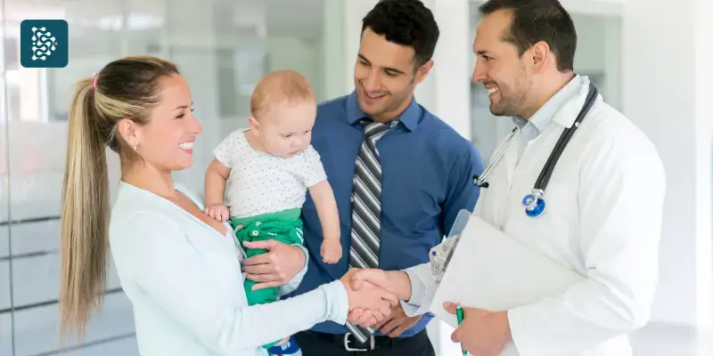 Família feliz com médica pediatra sorridente; mulher segura bebê no colo em ambiente de medicina humanizada.