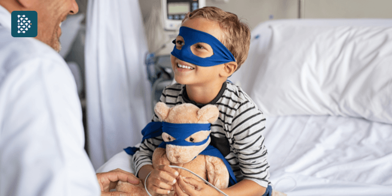 menino pequeno com máscara de super-heroi segurando ursinho conversando com médico pediatra no hospital infantil