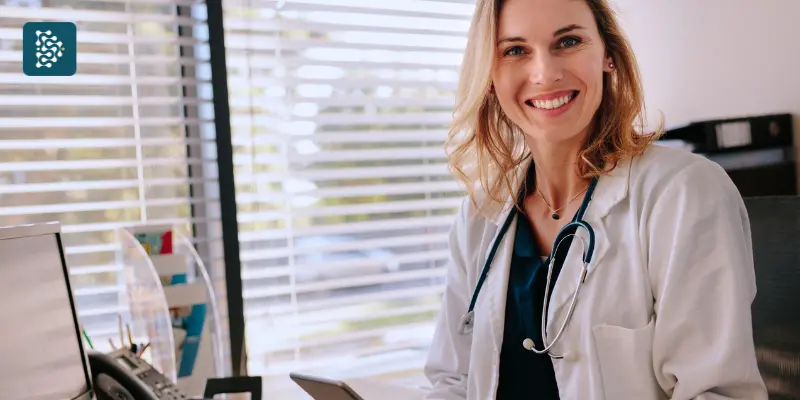 médica de braço cruzando sorrindo ao lado de uma maca em pose de liderança feminina na saúde usando sistema datasigh