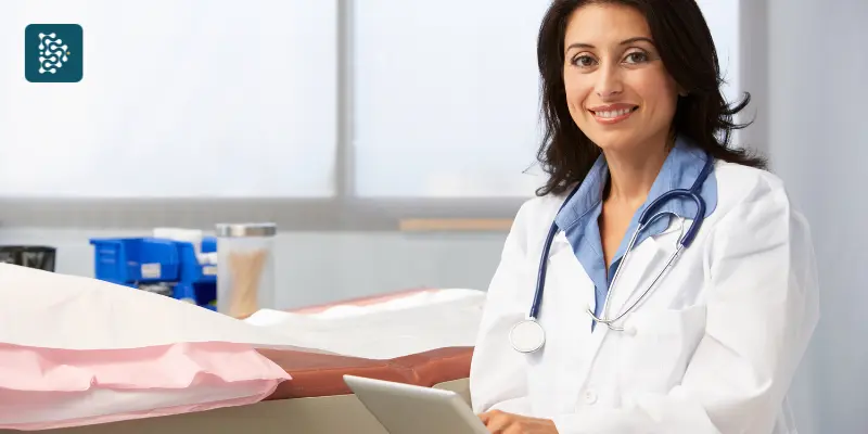 médica de braço cruzando sorrindo ao lado de uma maca em pose de liderança feminina na saúde usando sistema datasigh