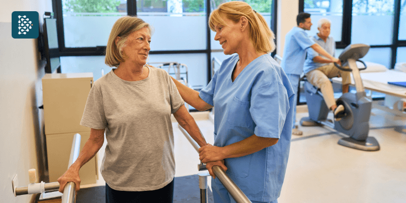 Lifetime Value na saúde. Mulher idosa amparada por enfermeira de jaleco azul durante sessão de reabilitação física no hospital, reforçando cuidado contínuo e fidelização do paciente.