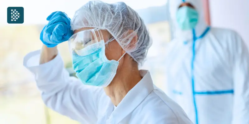 Médica preocupada com as mãos na cabeça, refletindo sobre sobrecarga de trabalho e cansaço nos hospitais.